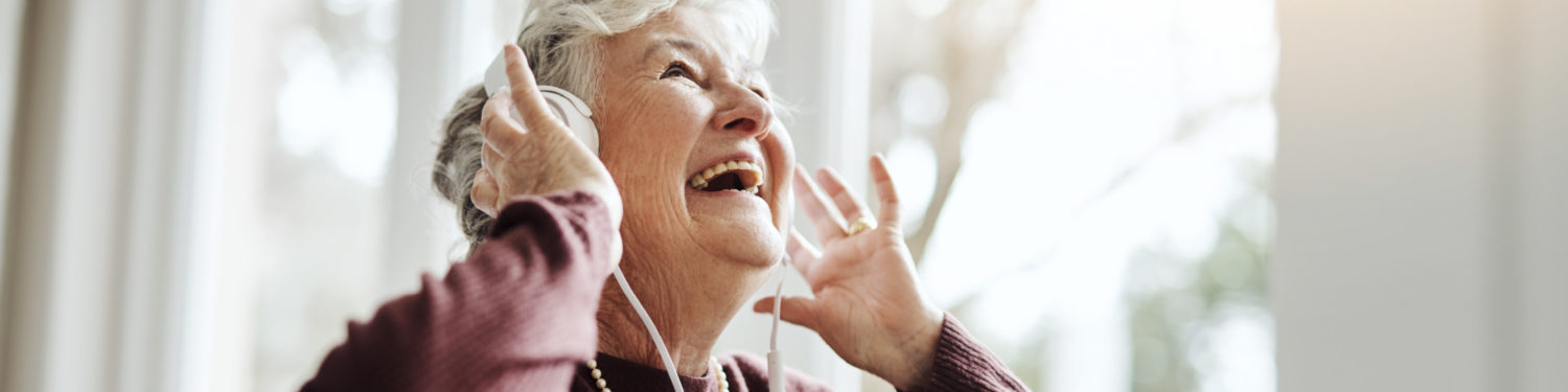 Iäkäs, hymyilevä, harmaahiuksinen nainen katsoo kuulokkeet korvillaan ylöspäin.