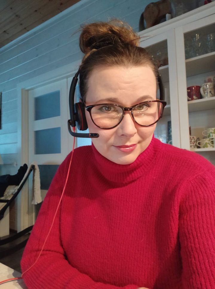 Silmälasipäinen nainen punaisessa paidassa istuu kotitoimistossaan kuulokkeet päässään.